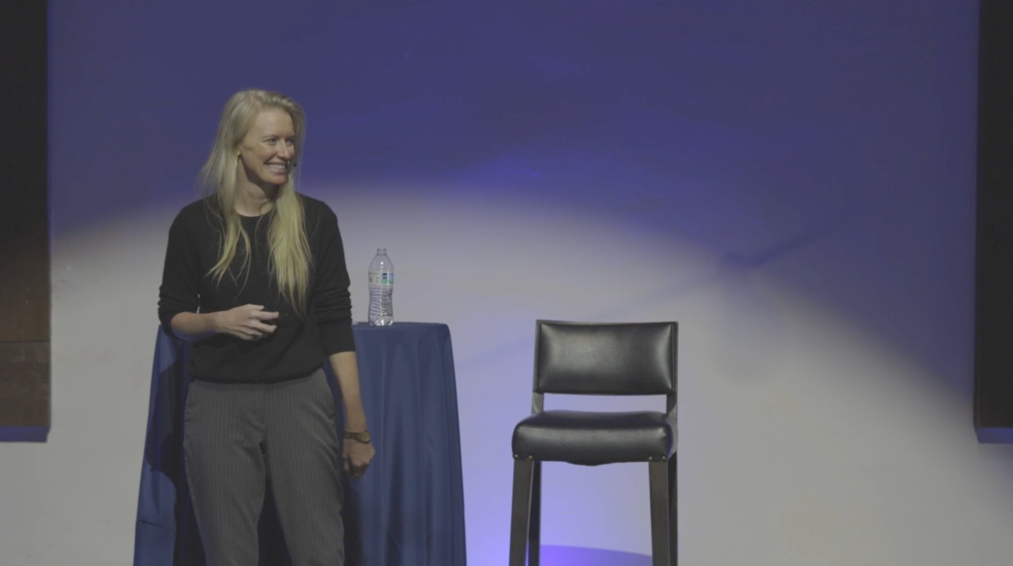 Bridget Hilton smiling on stage beside a cocktail table and stool