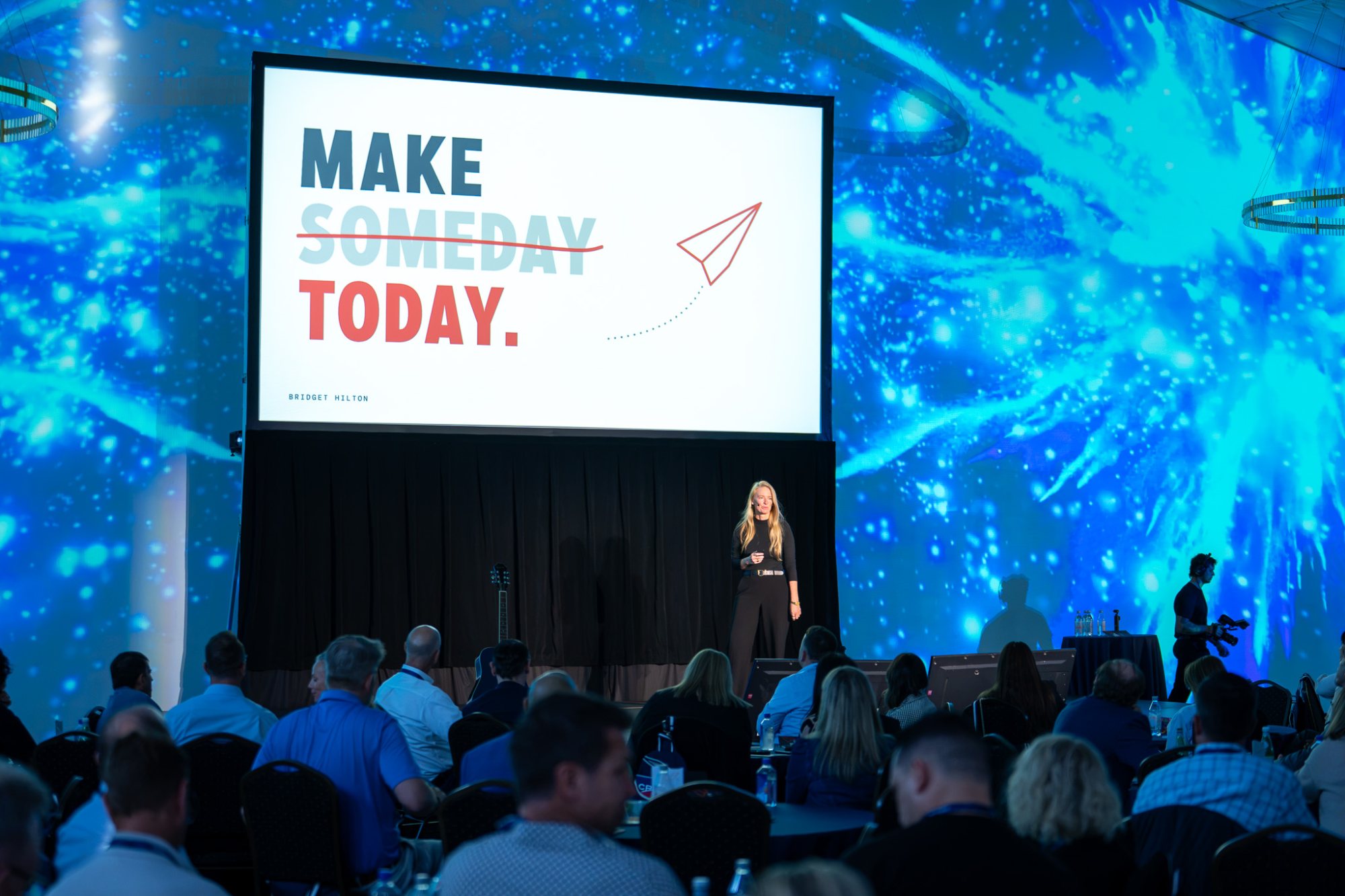 Bridget Hilton on stage beneath a giant Make Someday Today slide, blue projection wall behind her